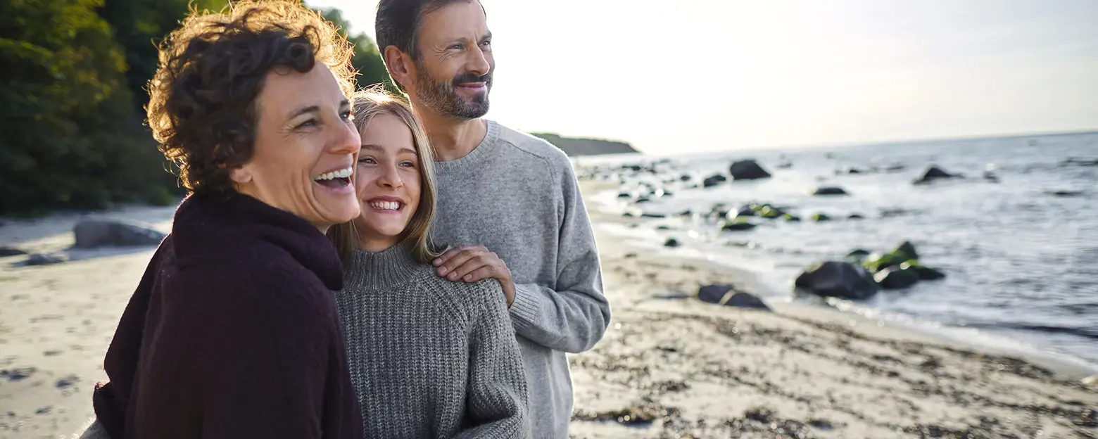 Trzy uśmiechnięte osoby, dwie dorosłe kobiety i jeden mężczyzna, stoją na plaży. Obejmują się w radosnej chwili, a w tle widać morze i kamienie. Słońce świeci, a atmosfera jest przyjemna i rodzinna.