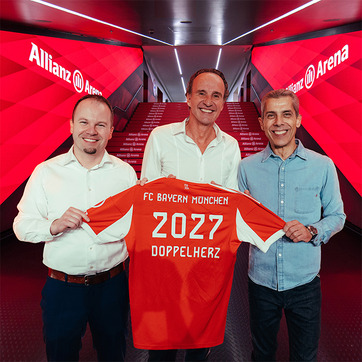 Trzech mężczyzn stoi w Allianz Arena, trzymając w rękach piłkarską koszulkę FC Bayern Monachium. Koszulka jest w kolorze pomarańczowym z napisem „2027” oraz „DOPPELAHRZ”. W tle widoczna jest schodnia prowadząca do stadionu.